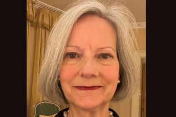 An older female with grey hair, facing the camera directly, wearing a pearl necklace and black blouse